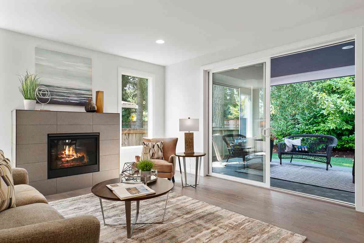 Cozy living room with fireplace, large windows overlooking backyard patio. Neutral tones, wooden accents.