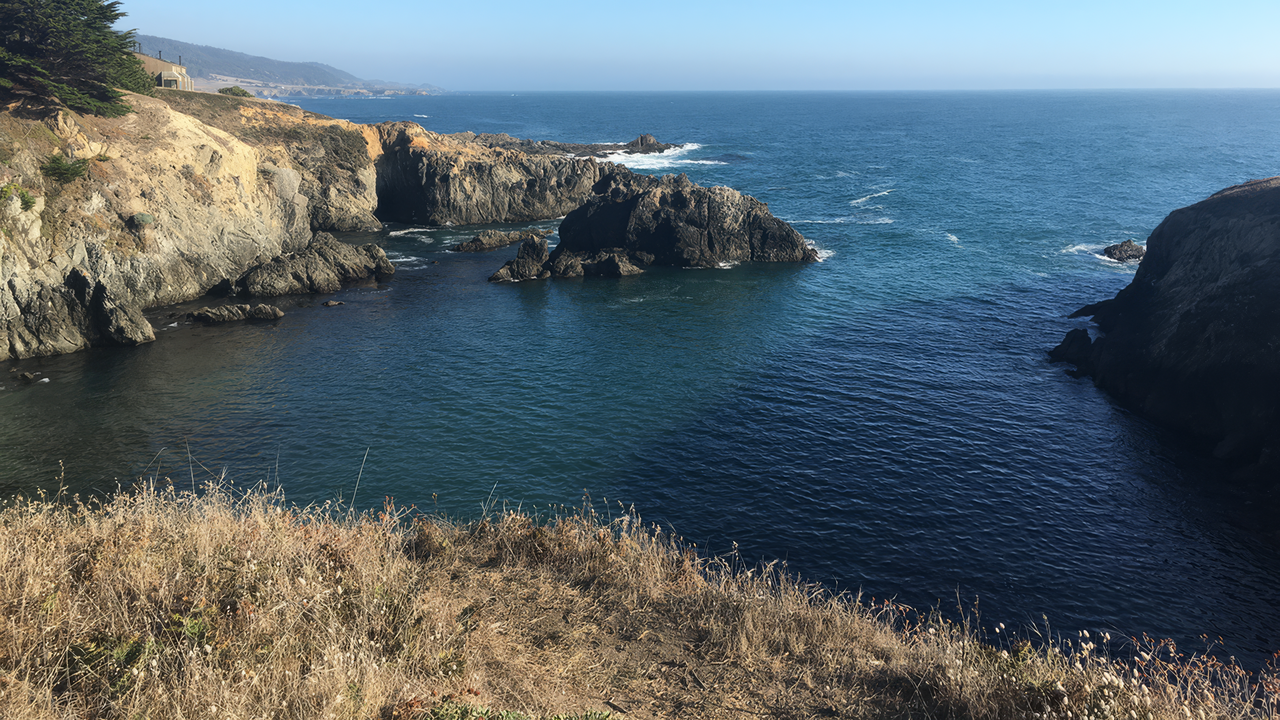 Rocky coastal landscape with rugged cliffs, blue ocean, and distant mountains.