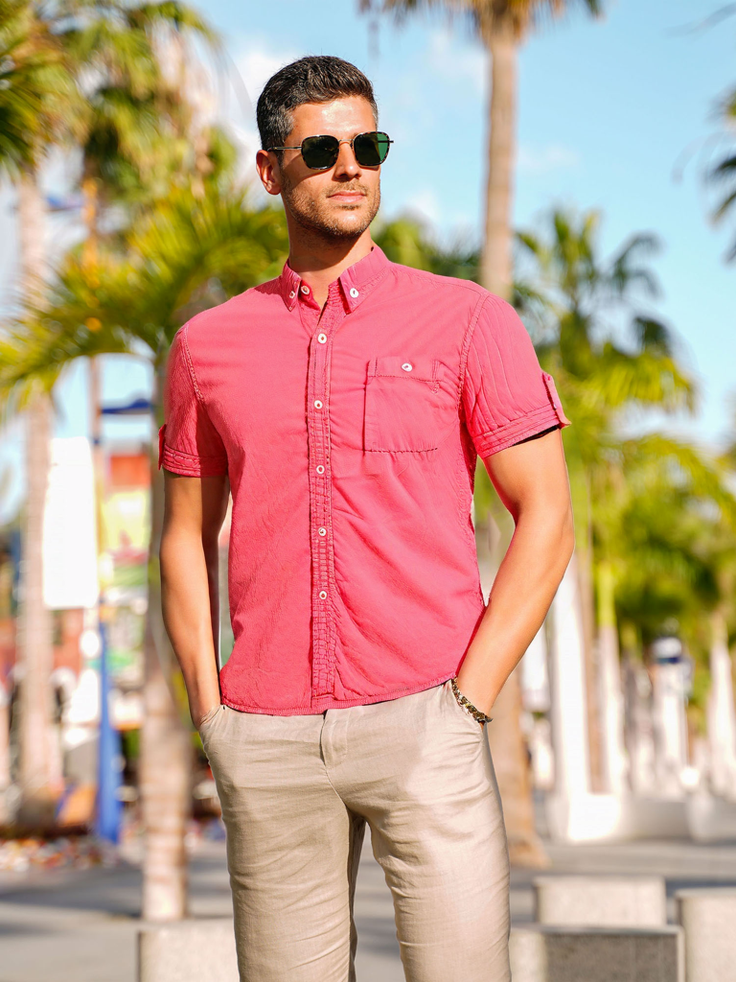 Um homem usando óculos escuros, olhando para a esquerda, vestindo uma camisa de botões rosa-avermelhada.